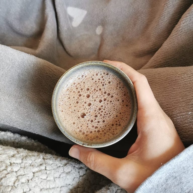 Eine Hand hält eine Tasse mit Ritualkakao auf einem grauen, weichen Untergrund.